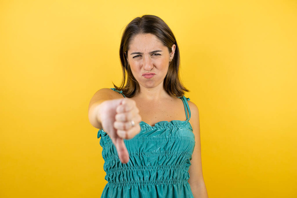 Молодая красивая женщина на изолированном желтом фоне с сердитым лицом, негативный знак показывает неприязнь с опущенным большим пальцем - Фото, изображение