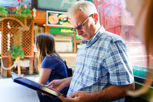 Seitenblick auf einen älteren Mann, der an sonnigen Sommer- oder Herbsttagen im Restaurant sitzt und die Speisekarte prüft, die zur Bestellung bereit ist - echtes Tourismuskonzept für Menschen - Foto, Bild
