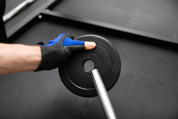 Male hand with a blue, gym glove loading weight plate on a chrome barbell, on a black, safe flooring mat. Bodybuilding in a fitness club, home gym. Sports equipment for weight training. - Фото, зображення
