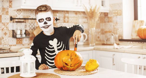 Onnellinen poika kotona keittiössä Halloween-puvuissa ja meikissä, veistää kurpitsan, leikkii ja nauraa. Valmistautuminen halloweeniin. - Valokuva, kuva