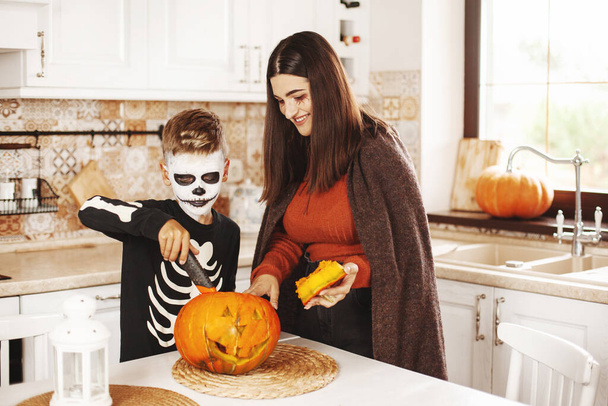 Bruder und Schwester sind in Halloween-Kostümen und -Schminke in der Küche zu Hause, schnitzen einen Kürbis, spielen und lachen. - Foto, Bild