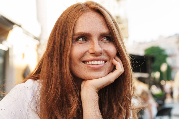 Close up of beautiful smiling red haired woman sitting at the cafe outdoors, looking away - 写真・画像