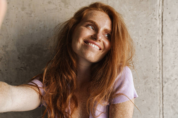 Image of redhead cheerful woman smiling and taking selfie photo while posing near concrete wall at home - Fotó, kép