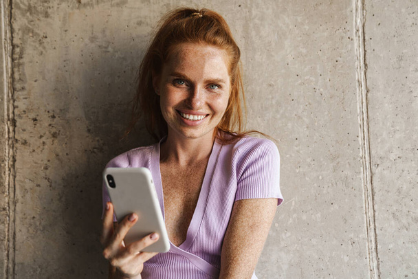 Image of redhead joyful woman smiling and using cellphone while standing near concrete wall at home - Fotografie, Obrázek