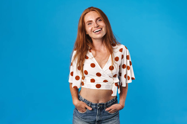 Portrait of a beautiful cheerful young woman with long red hair wearing summer clothes standing isolated over blue background, looking at camera - Фото, изображение