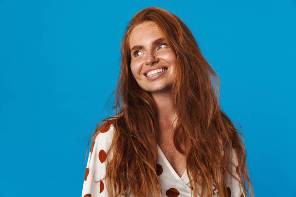 Portrait of a beautiful cheerful young woman with long red hair wearing summer clothes standing isolated over blue background, looking away - Fotó, kép