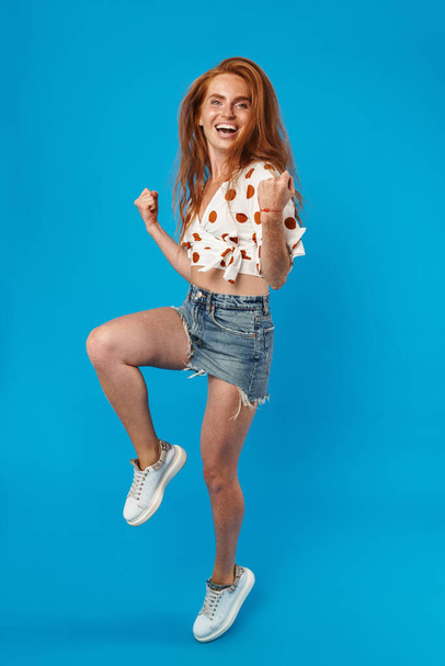 Full length portrait of a beautiful cheerful young woman with long red hair wearing summer clothes jumping isolated over blue background, celebrating - Фото, зображення