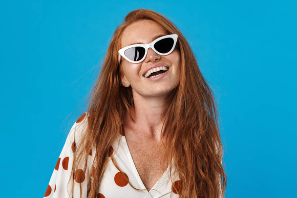 Portrait of a beautiful cheerful young woman with long red hair wearing summer clothes and sunglasses standing isolated over blue background - Zdjęcie, obraz
