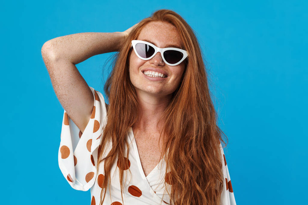 Portrait of a beautiful cheerful young woman with long red hair wearing summer clothes and sunglasses standing isolated over blue background, posing - Фото, изображение