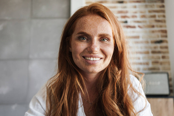 Image of happy ginger woman with freckles smiling and looking at camera in home - Photo, image