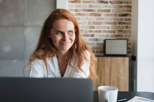 Image of smiling redhead businesswoman working with laptop while sitting at table in home - Foto, afbeelding