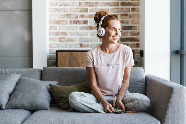 Obrázek usmívající se mladé ženy pomocí sluchátek a mobilu při sezení na gauči doma - Fotografie, Obrázek