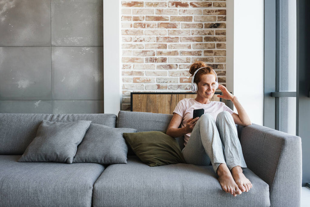 Image of smiling young woman using headphones and cellphone while sitting on couch at home - Фото, изображение