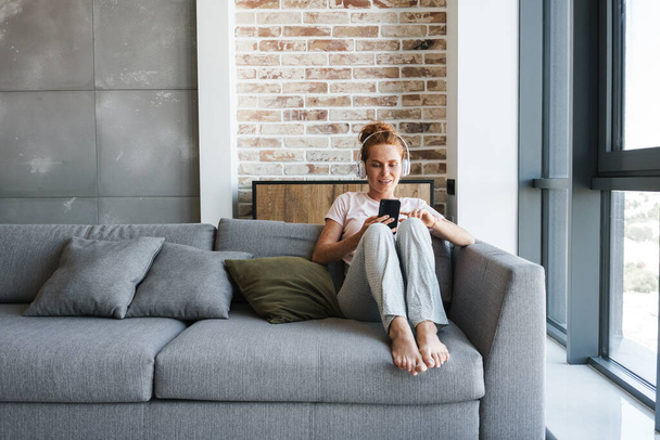 Image of pleased young woman using headphones and cellphone while sitting on couch at home - Foto, Bild