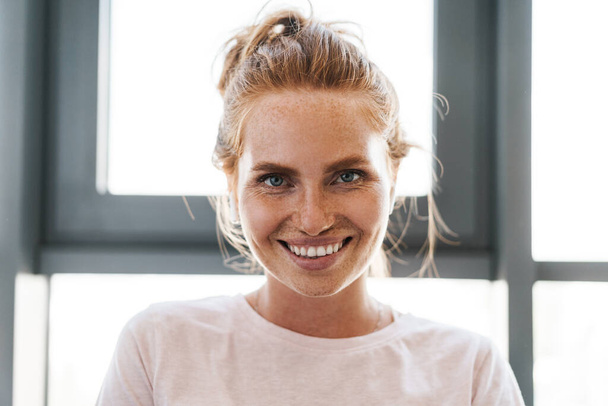 Image of happy ginger woman with freckles smiling and using wireless earphones in home - Foto, afbeelding