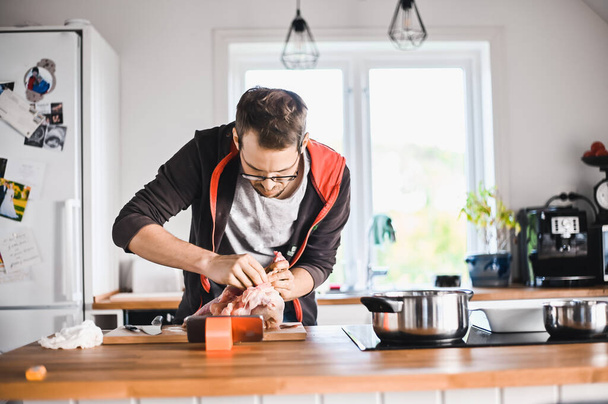 atractivo chico nerd con gafas en la cocina con estilo sigue una receta en el teléfono prepara pollo - Foto, Imagen