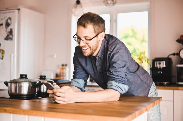 attractive nerd guy with glasses in stylish kitchen follows a recipe or  reads news on the phone - Foto, Imagen