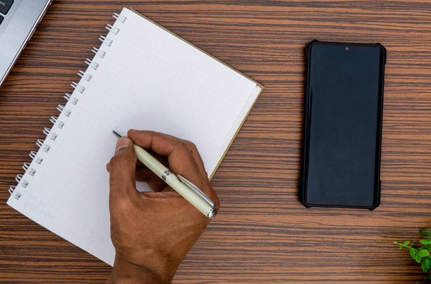 Írni egy jegyzetfüzetre, miközben otthonról dolgozol. Ezen a barna csíkos munkaasztalon egy körtető, egy mobil, egy számológép és egy növény is látható.. - Fotó, kép