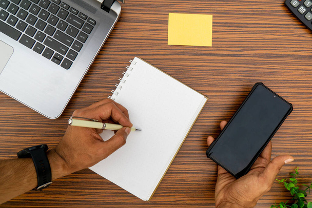 Írni egy jegyzettömbre, miközben egy mobiltelefont tart irodai környezetben. Ezen a barna csíkos munkaasztalon egy körtető, egy mobil, egy számológép és egy növény is látható.. - Fotó, kép