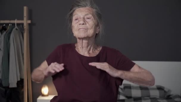 Bardzo stara kobieta z siwymi włosami i głębokimi zmarszczkami wykonuje ćwiczenia fizyczne w domu. Starsza kobieta rozgrzewa się w małym mieszkaniu. Happy senior fitness w drzwiach podczas covid 19 - Materiał filmowy, wideo