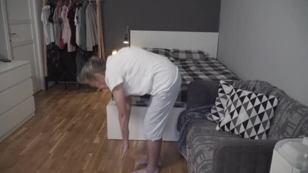 Very old age woman with gray hair and deep wrinkles does physical exercises at home. An elderly female is warming up in a small apartment. Happy senior exercising fitness within doors during covid 19 - Video, Çekim