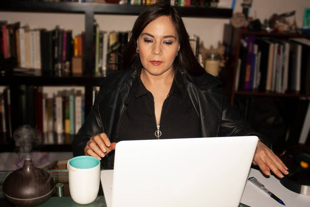 Žena ve věku 40-45 let pracující v domácí kanceláři příjemně doprovází kávu a zvlhčovač s párou a vůní, která zkrášluje životní prostředí za ní je velká knihovna s podpůrnými knihami v Mexico City - Fotografie, Obrázek