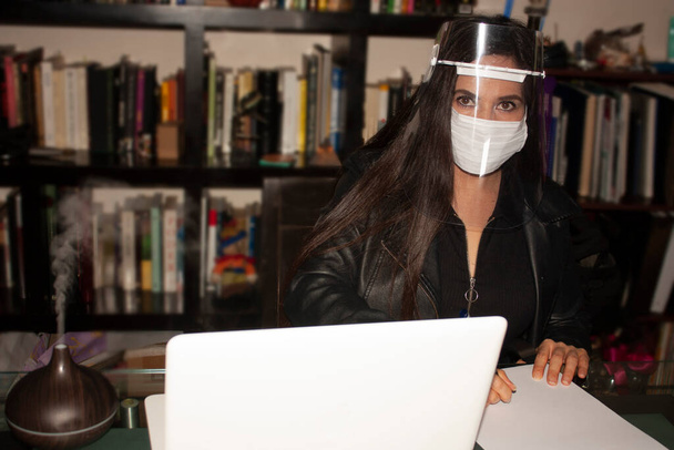 Mexicaanse vrouw leeft de nieuwe rallity met inachtneming van normen voor covid-19 Pandemie in Mexico stad met een masker en masker of mond covers in het kantoor met een boekenkast op de achtergrond, terwijl het schrijven van notities en ademlucht en luchtbevochtiger damp te verbeteren t - Foto, afbeelding