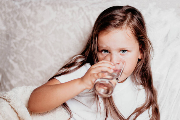 un bambino malato, una ragazza seduta su un letto con un cuscino bianco, che beve acqua da un bicchiere - Foto, immagini