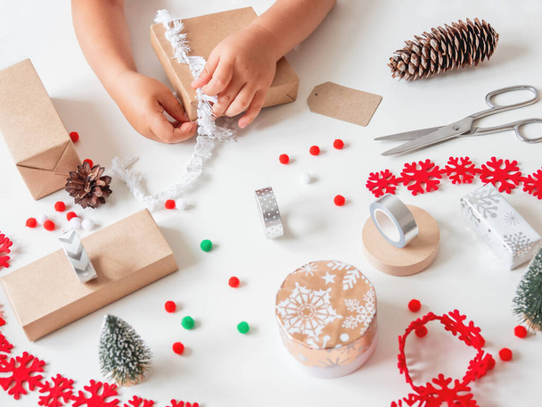 Kid wraps handmade Christmas presents in craft paper with colorful pompons and snowflake ribbons. Child prepares gifts for New Year celebration. Peaceful leisure activity before winter holidays. - Фото, изображение