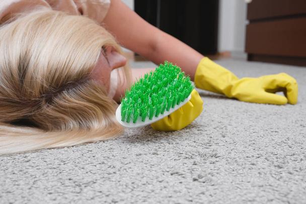 Woman in yellow gloves with a green brush is so tired of cleaning and brushing carpet, removing stains and wool from it and doing routine homework, that she lay down on the floor and sleep - Foto, Imagen