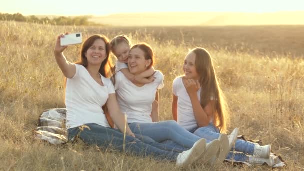 Mutlu aile seyahatleri, parkta akıllı telefondan selfie. Anne ve sağlıklı çocuklar battaniyenin üzerinde oturuyorlar. Anne ve kızları battaniyeyle tarlada fotoğraf çekiliyor. Aile seyahati ve macera - Video, Çekim