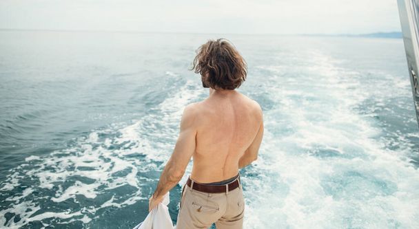 Вид сзади на молодого человека, сидящего на краю яхты, смотрящего на море - Фото, изображение