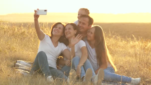 Onnellinen perhe matkustaa. selfie älypuhelimessa puistossa. Äiti, isä ja lapset istuvat peiton päällä kuvaamassa videota. Äiti ja isä tyttärineen kuvataan pellolla. perhe bloggaajia - Materiaali, video