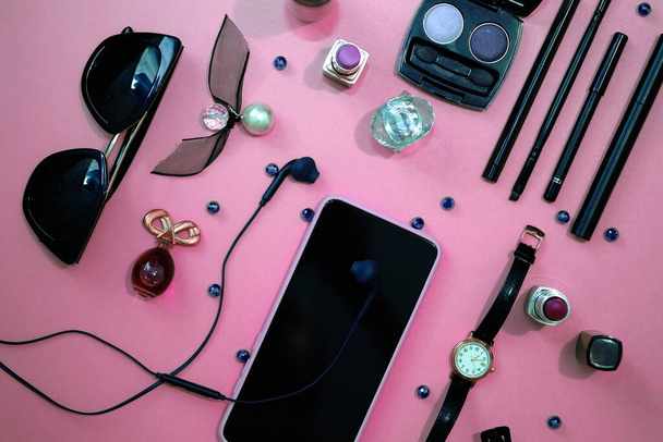 女性の化粧品やアクセサリーのトップビュー。ピンクの背景に電話、時計、ヘッドフォン、化粧品の構成。女性の財布の中にあるもの. - 写真・画像