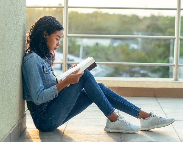 Egy ázsiai arcvonású főiskolás lány könyvet olvas a padlón ülve. - Fotó, kép