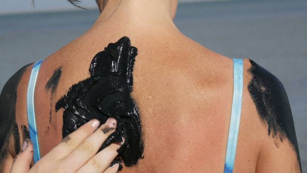 Healing black medical mud applies on back woman at the lake of salt water. Salty black dirt Is good for people with health and skin problems. Relax at spa resort. Exfoliation scrub beauty treatment - Photo, Image