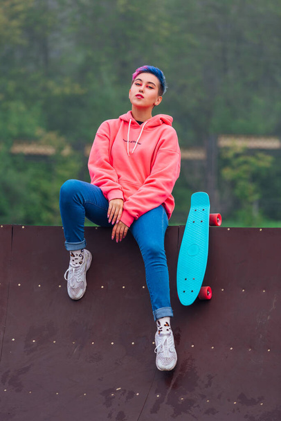 Young girl with short multicolored hair sits with skateboard in skatepark - Fotoğraf, Görsel
