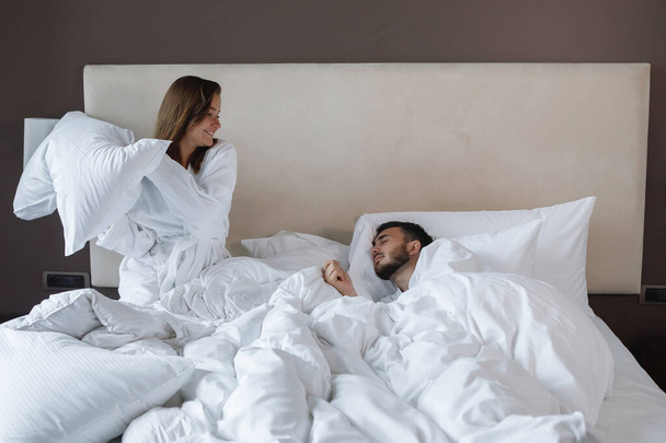 Woman in a white bed wakes a sleeping bearded man with a pillow. Rest in the hotel, mating games. - Foto, Imagem