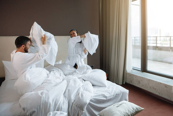 白いベッドの女と男が大きな窓で寝室で枕と戦っている。ホテルで休んで、ゲームを交配. - 写真・画像