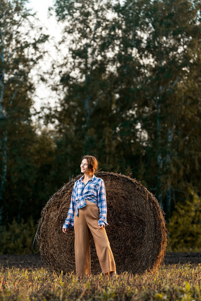 Junge schöne brünette Frau in beiger Hose und Hemd posiert vor dem Hintergrund von Heuhaufen in einem geschnittenen Feld - Foto, Bild