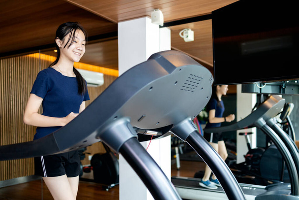 Sağlıklı bir Asyalı kız çocuğu sağlık için koşu pisti kullanıyor, egzersiz makinelerinde koşmadan önce ısınmak için koşu bandında yürüyen insanlar, yanında büyük bir ayna, spor salonunda spor yapıyor, sağlık hizmetlerinde çalışıyor. - Fotoğraf, Görsel