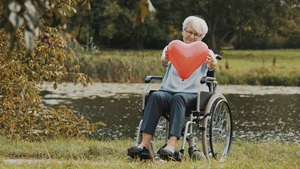 mulher idosa feliz na cadeira de rodas perto do rio oferecendo um coração de balão com um sorriso. Conceito de amor e cuidado - Foto, Imagem