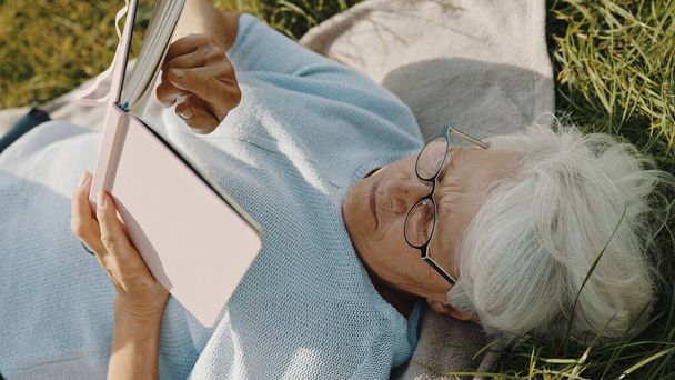 starší šedovlasá žena čtení knihy, zatímco odpočívá na trávě v přírodě - Fotografie, Obrázek