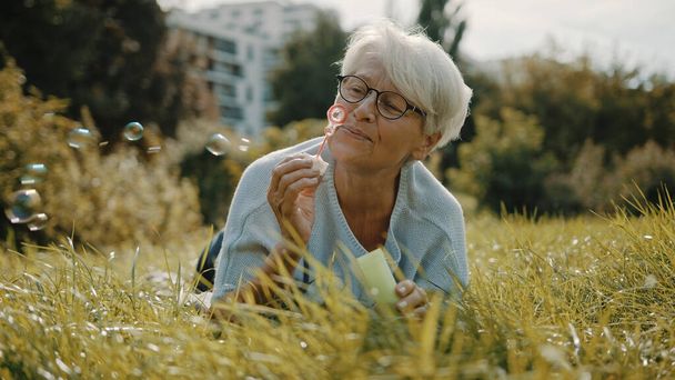 Retired senior woman enjoying freedom. Childish grandma blowing soap bubbles in the park - Photo, Image