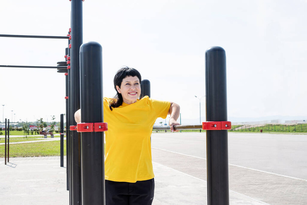 Smiling senior woman doing reverse push ups outdoors on the sports ground bars - Photo, Image