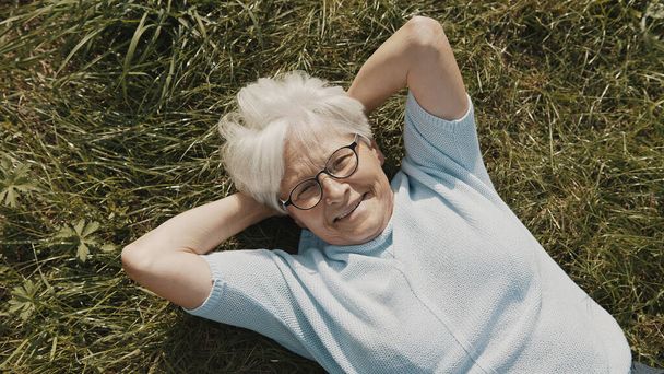 ηλικιωμένη γυναίκα, συνταξιούχος ξαπλωμένη στο γρασίδι με τα χέρια κάτω από το κεφάλι της στον κήπο από πάνω κοντινό πλάνο. - Φωτογραφία, εικόνα
