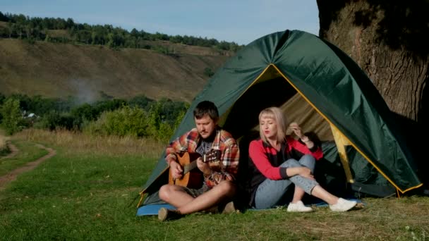 Man and woman sing a song with guitar sitting at tent. Family is on camping trip. - Footage, Video