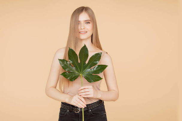 Beautiful blonde girl with clean skin holds green leaf on beige background. Beauty spa care cream concept - Photo, Image