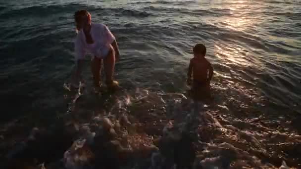 Νεαρή γυναίκα και ο γιος της έρχονται στο νερό το ηλιοβασίλεμα στην αμμώδη παραλία, διακοπές στο νησί - Πλάνα, βίντεο