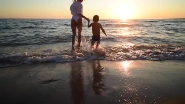 Mujer joven y su hijo vienen al agua en la puesta de sol en la playa de arena, vacaciones en la isla - Imágenes, Vídeo